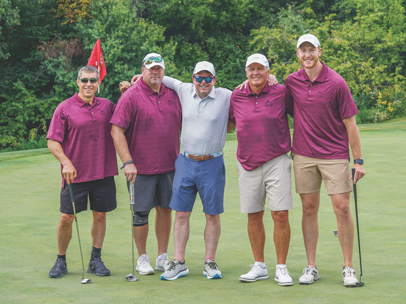 Five golfers pose together on a putting green; four wear matching maroon polos.